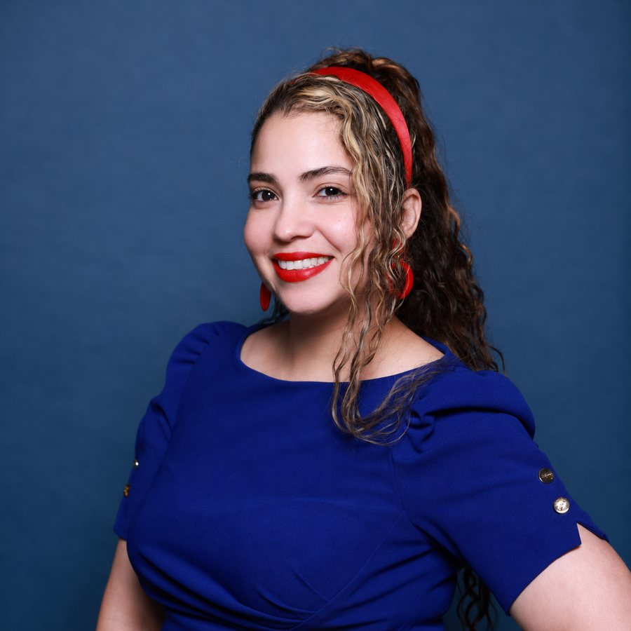 Nayeli Castillo smiling straight at the camera in a cobalt blue dress and red headband against a deep indigo studio background.