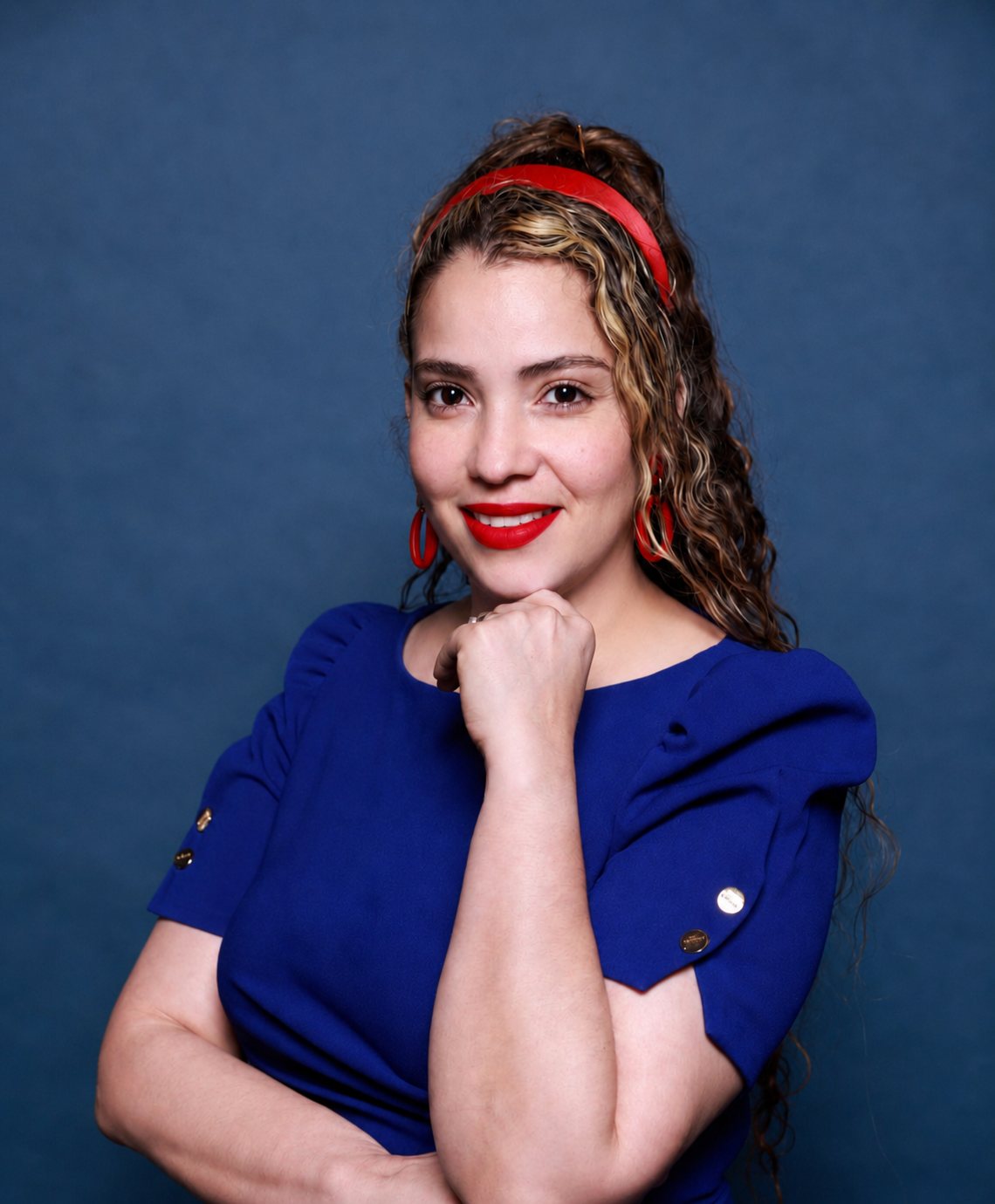 Nayeli Castillo, chin resting on her hand, looking directly at the camera — cobalt blue dress and red headband against a deep indigo studio background.